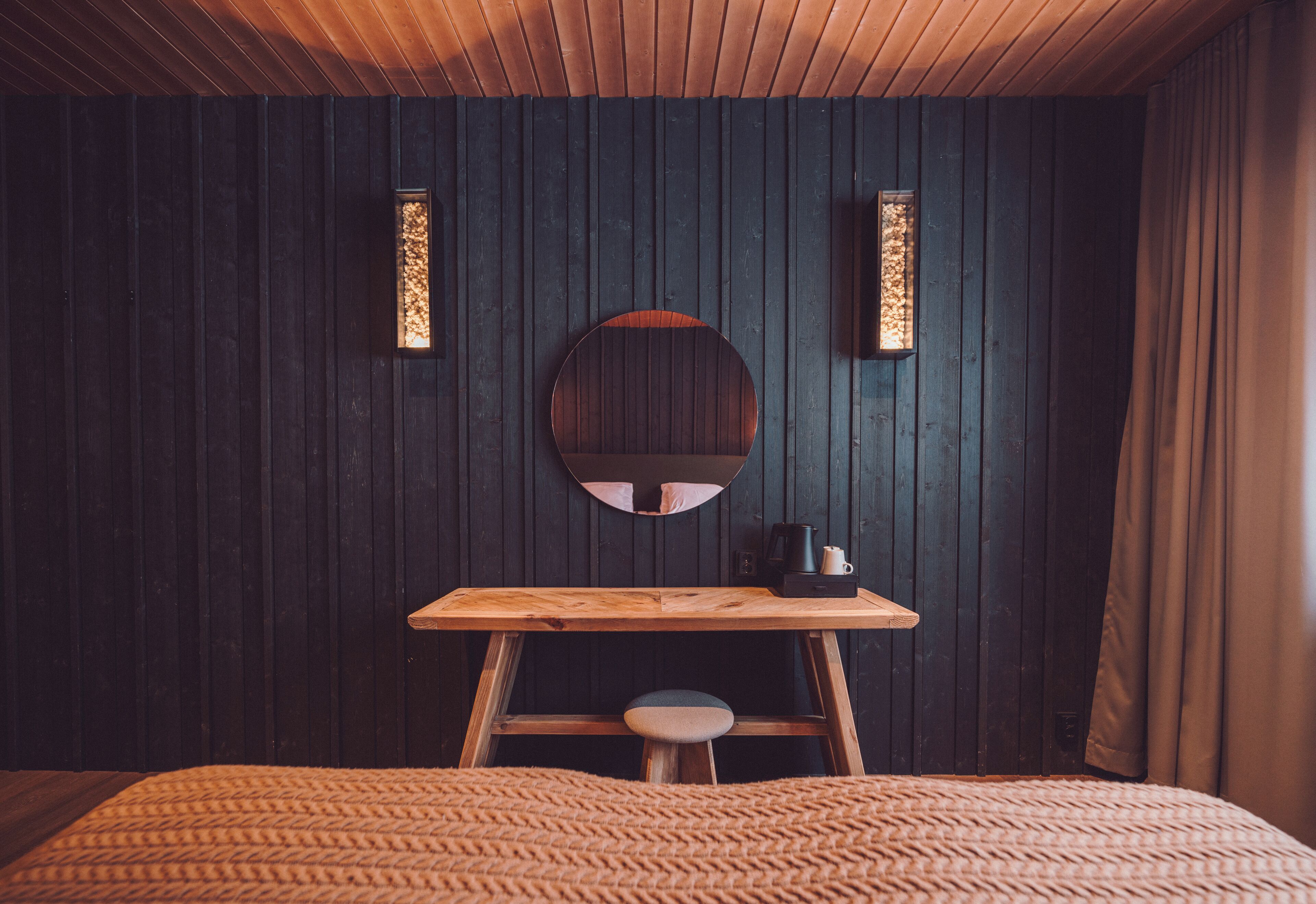 A cozy room with a wooden bed, a small stool, a round mirror, and two wall-mounted lights.
