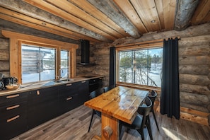 A log cabin interior with a wooden dining table, black cabinets, and large windows offering a view of the outdoors.
