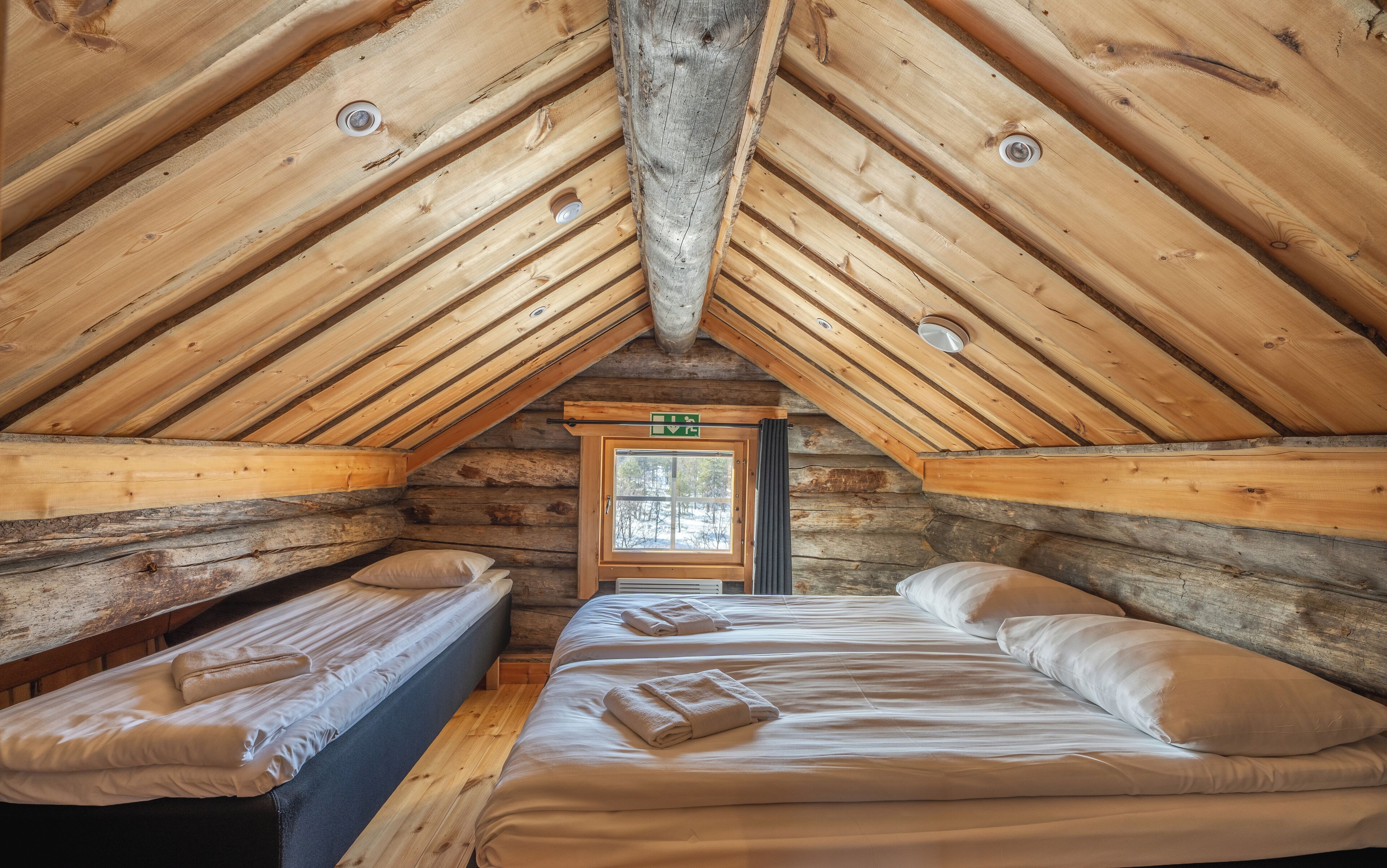 A log cabin with a sloped ceiling, two beds with towels, a window, and a door with an exit sign.