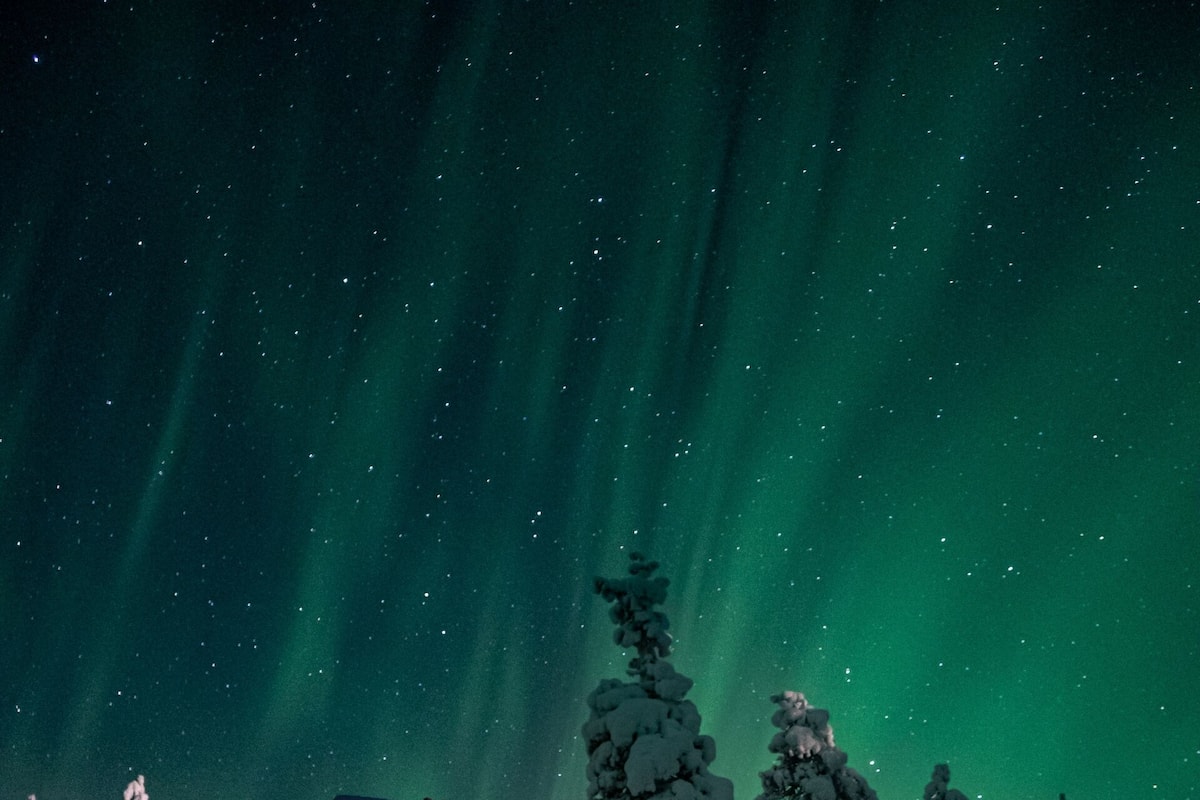 Une cabane en rondins, dont l’intérieur est illuminé d’une lumière chaleureuse, entourée de neige et baignée sous un ciel étoilé.