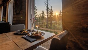 Een houten eettafel met borden eten en een glas wijn, met op de achtergrond een bos en een zonsondergang door een raam.