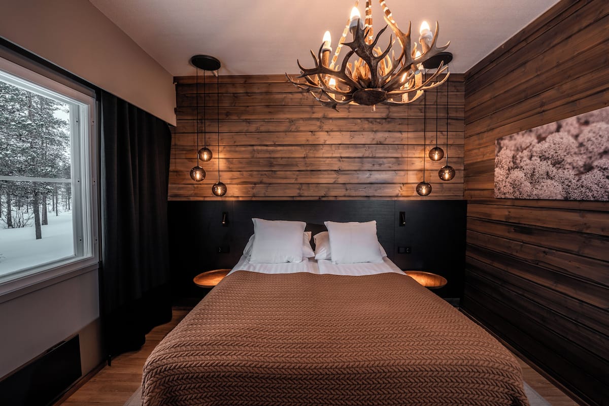 Une chambre à coucher avec une tête de lit en bois, un grand lit recouvert de draps blancs, un lustre et une fenêtre avec des rideaux.