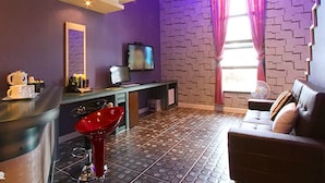 Una habitación con pared de ladrillo morado, un sofá moderno, un sillón rojo, una mesita con una tetera y un televisor en la pared.