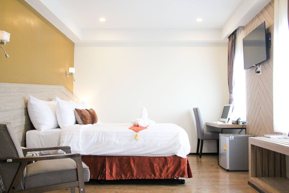 A hotel room with a bed, a desk with a computer, a chair, and a TV mounted on the wall.