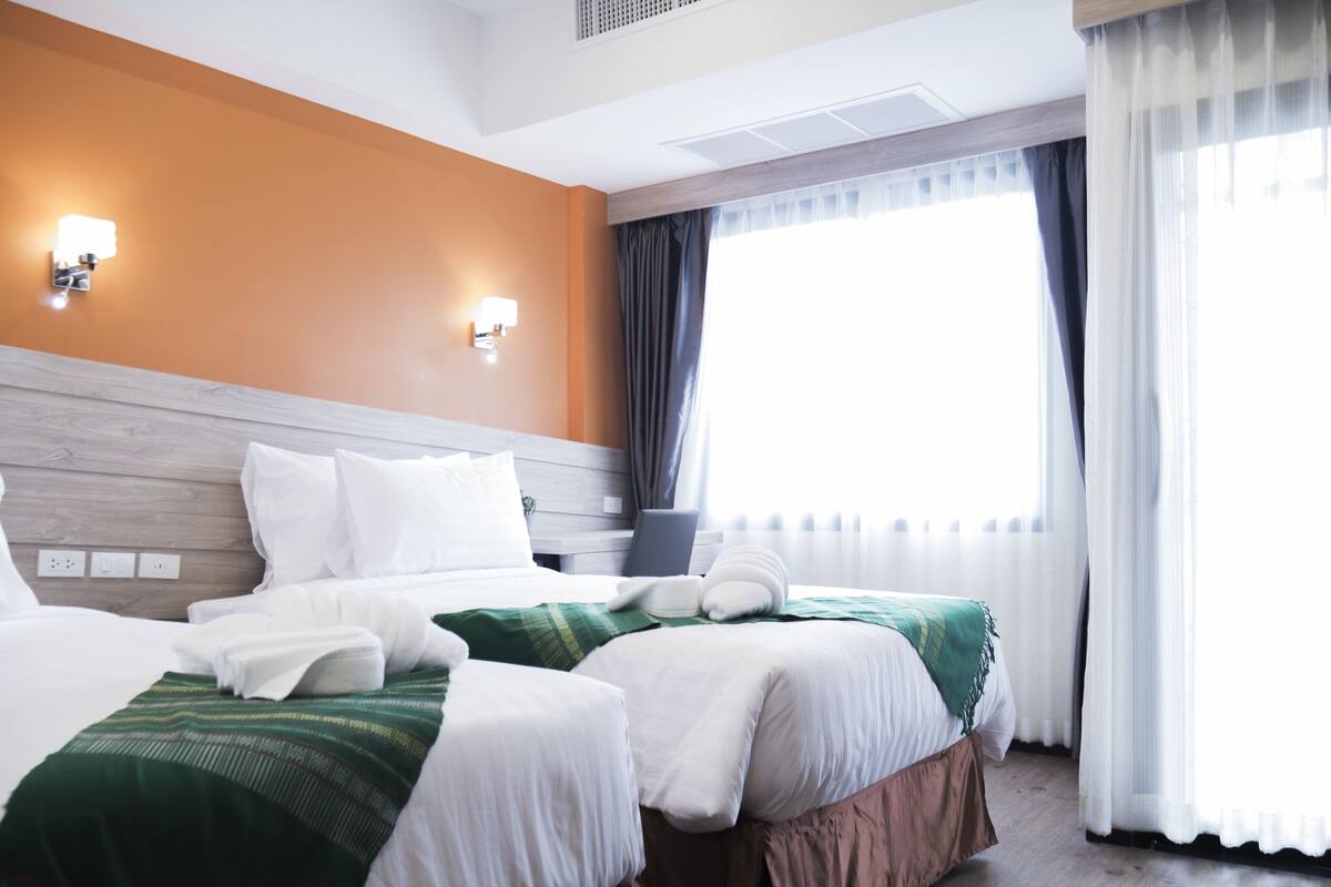 A hotel room with two beds, a wooden headboard, a large window with curtains, and wall-mounted lights.