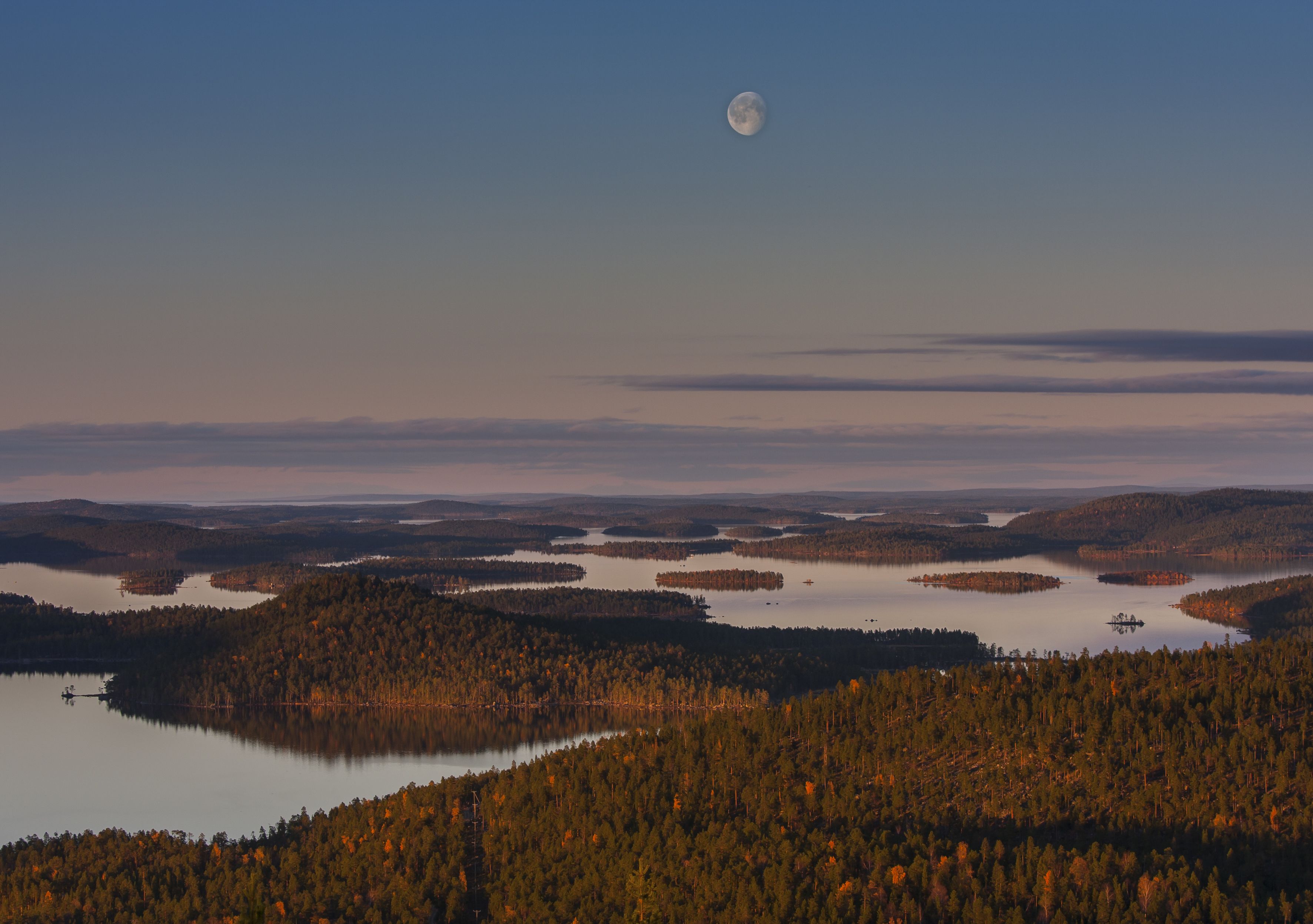 Un paisaje con una extensa masa de agua rodeada de colinas boscosas y una luna visible en el cielo.
