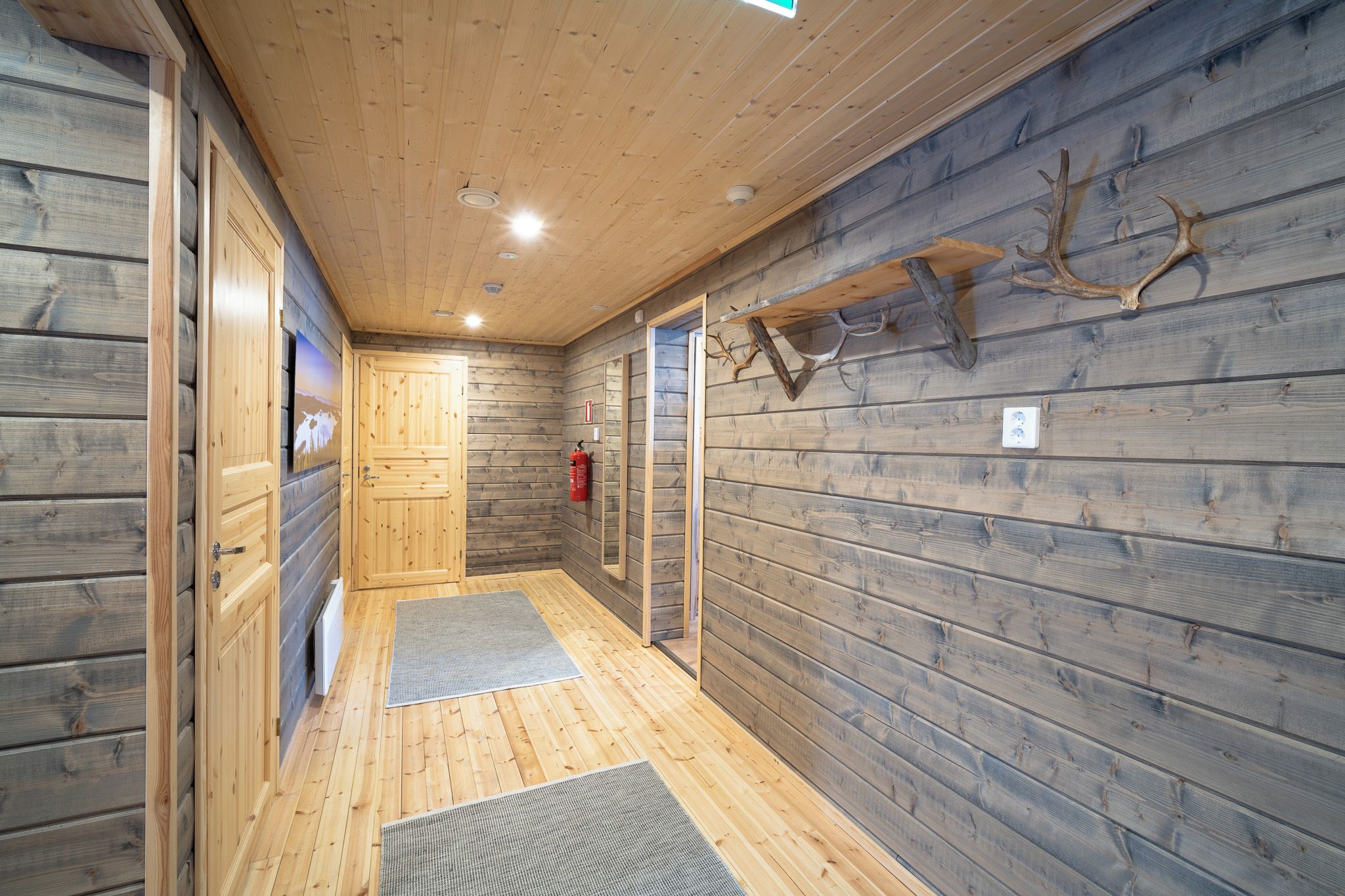 A wooden interior hallway with a fire extinguisher, a door, and a wall-mounted light fixture.
