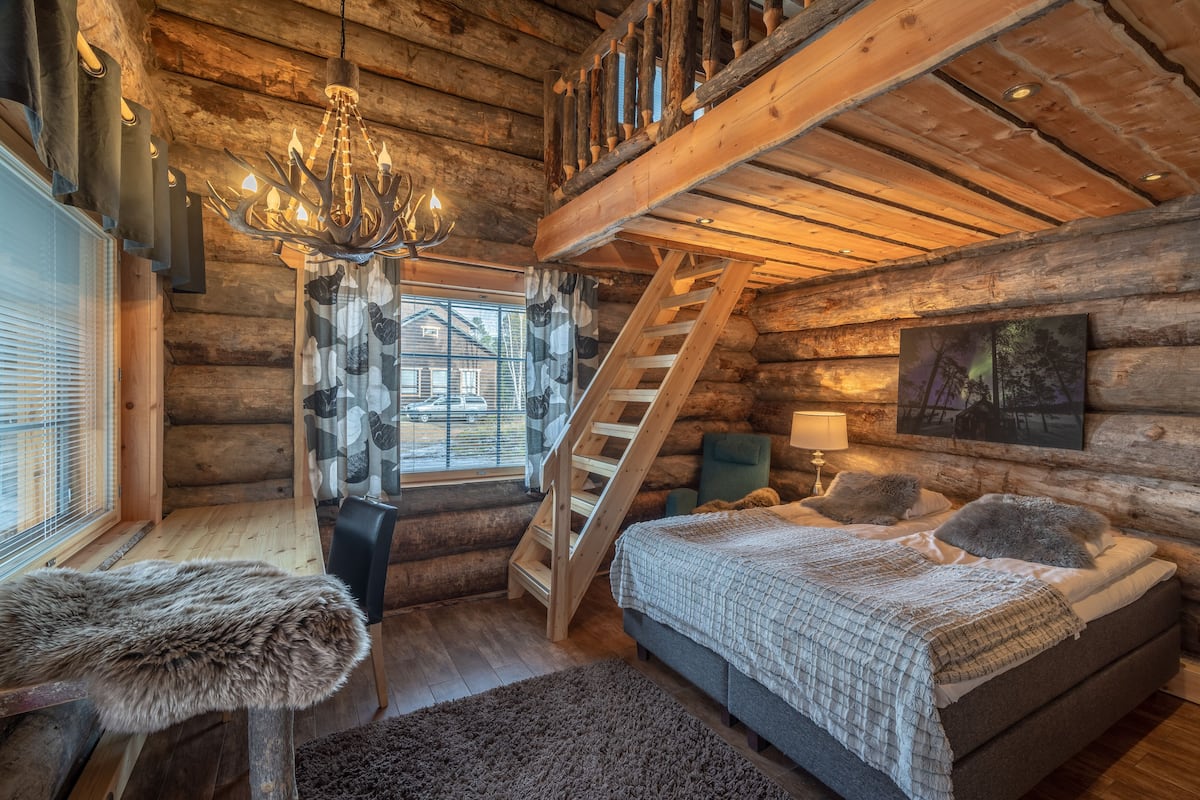 Un dormitorio tipo cabaña de madera con cama, candelabro, escalera y ventana con persianas.