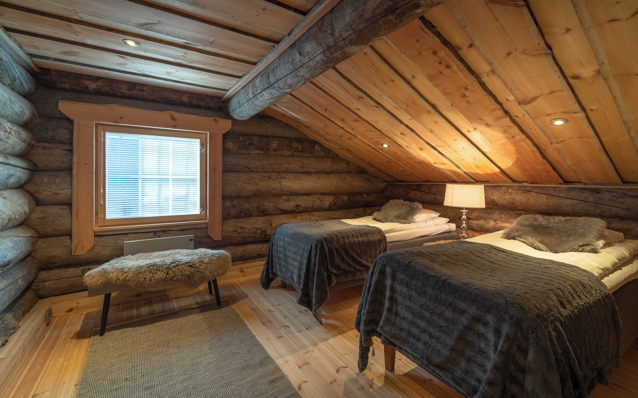 Un dormitorio tipo cabaña de madera con dos camas, una ventana con persianas, una lámpara y un banco cubierto de piel.