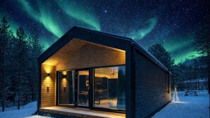 Uma cabana moderna com uma grande porta de vidro, fachada de madeira e vista do céu noturno com aurora boreal.