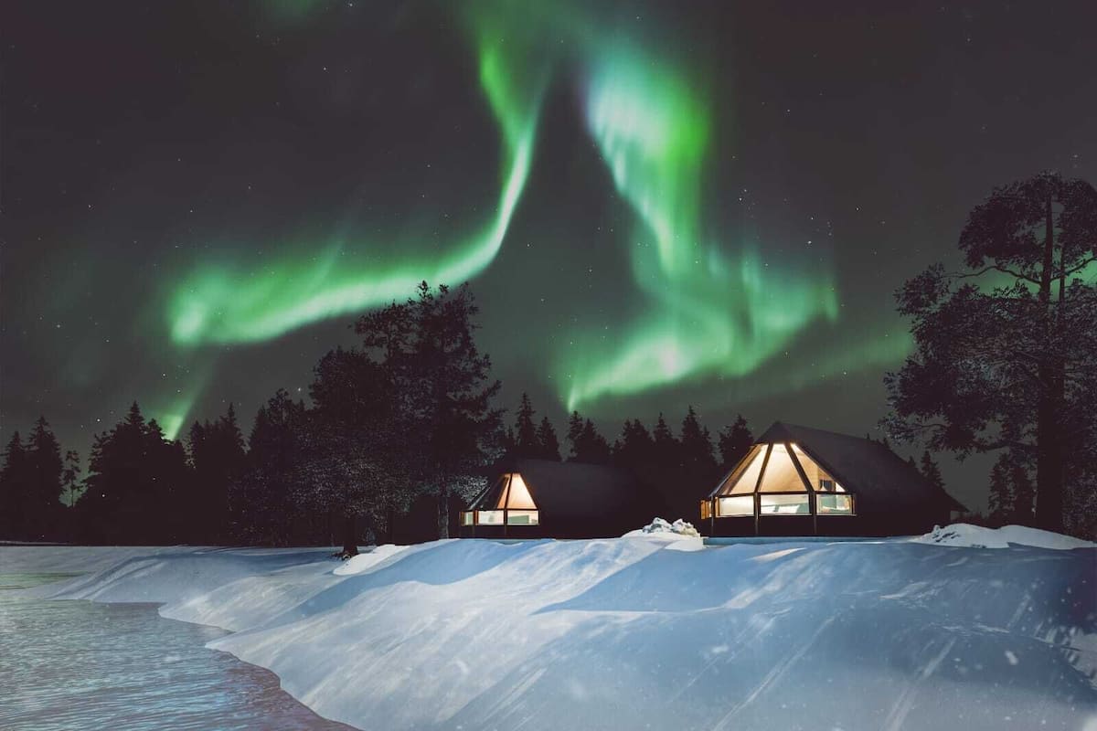 Una cabaña con fachada de vidrio, rodeada de terreno y árboles nevados, bajo un cielo nocturno con aurora boreal verde.