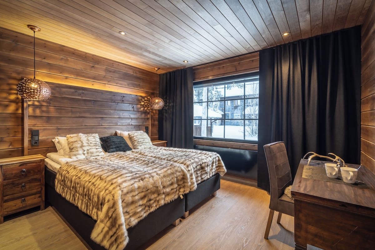 Un dormitorio acogedor con techo de madera, una cama grande con una manta de piel sintética, una mesita de noche, un escritorio con dos tazas y una ventana con cortinas.