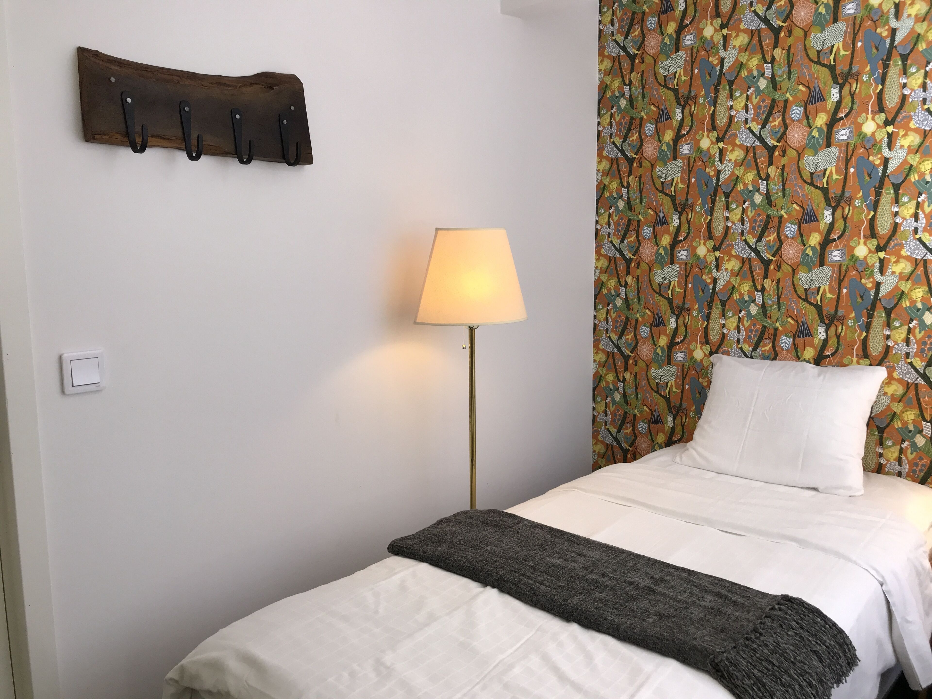 Una cama bien hecha con edredón blanco y manta gris, un perchero de madera fijado a la pared con ganchos, una lámpara de pie y un papel tapiz estampado.