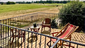 Un balcon donnant sur un champ verdoyant et des arbres. On y trouve deux fauteuils en osier et une petite table sur laquelle est posée une plante en pot.