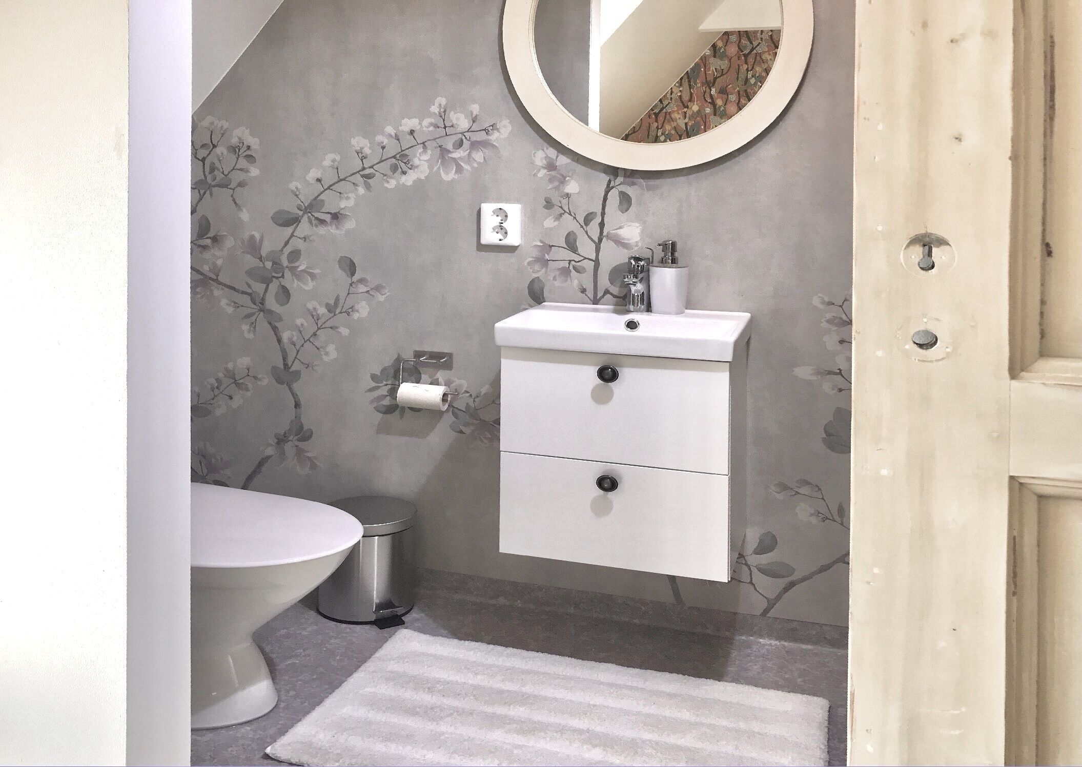A bathroom with a toilet, a sink with a mirror, a trash bin, and a patterned wall.
