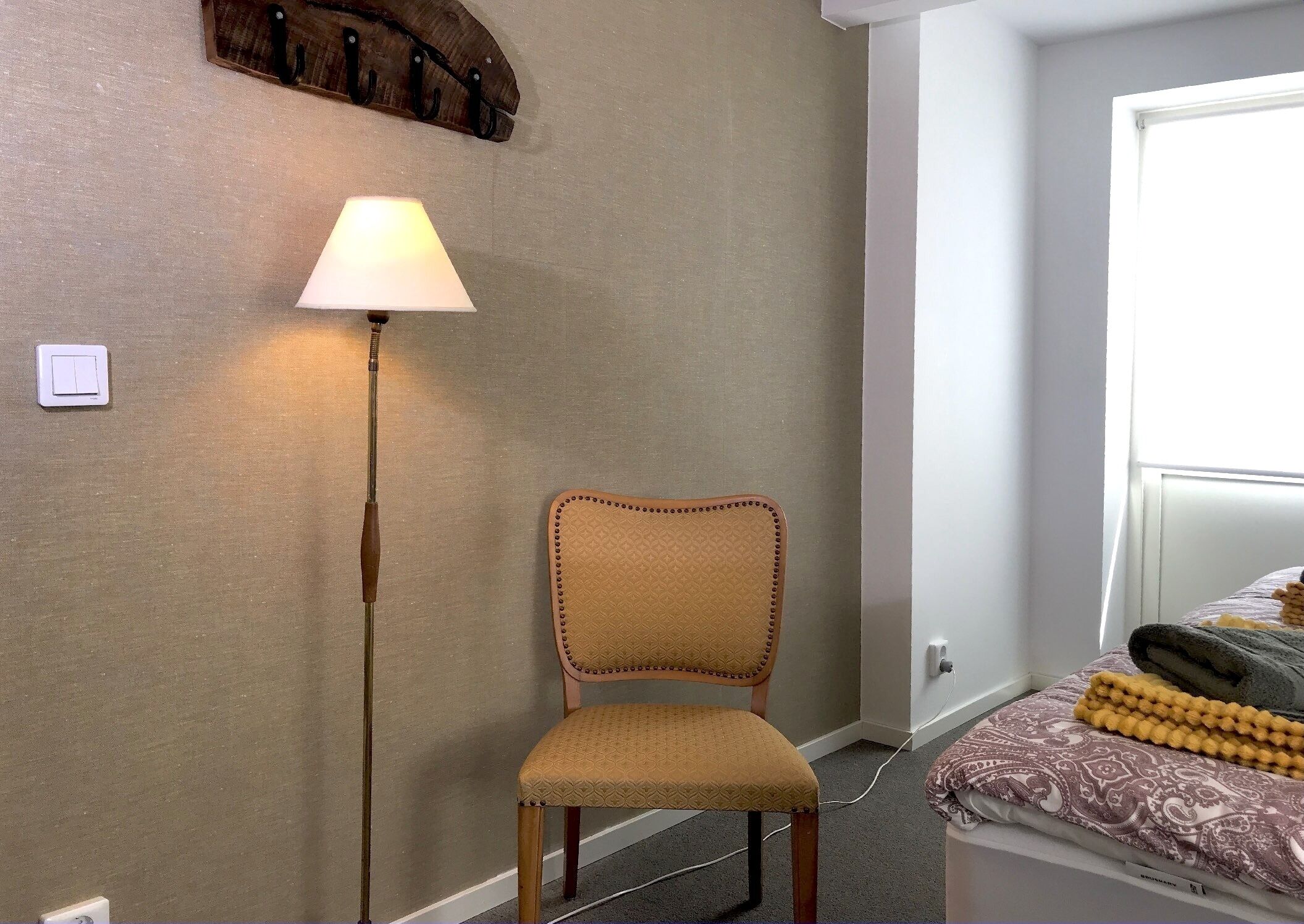 Una habitación con una silla, una lámpara de pie, una cama con ropa de cama estampada y una lámpara de pared.
