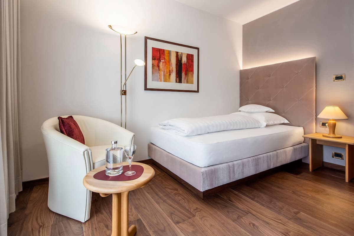 A hotel room with a bed, a chair, a small table with a glass of water, a lamp, and a framed abstract painting on the wall.