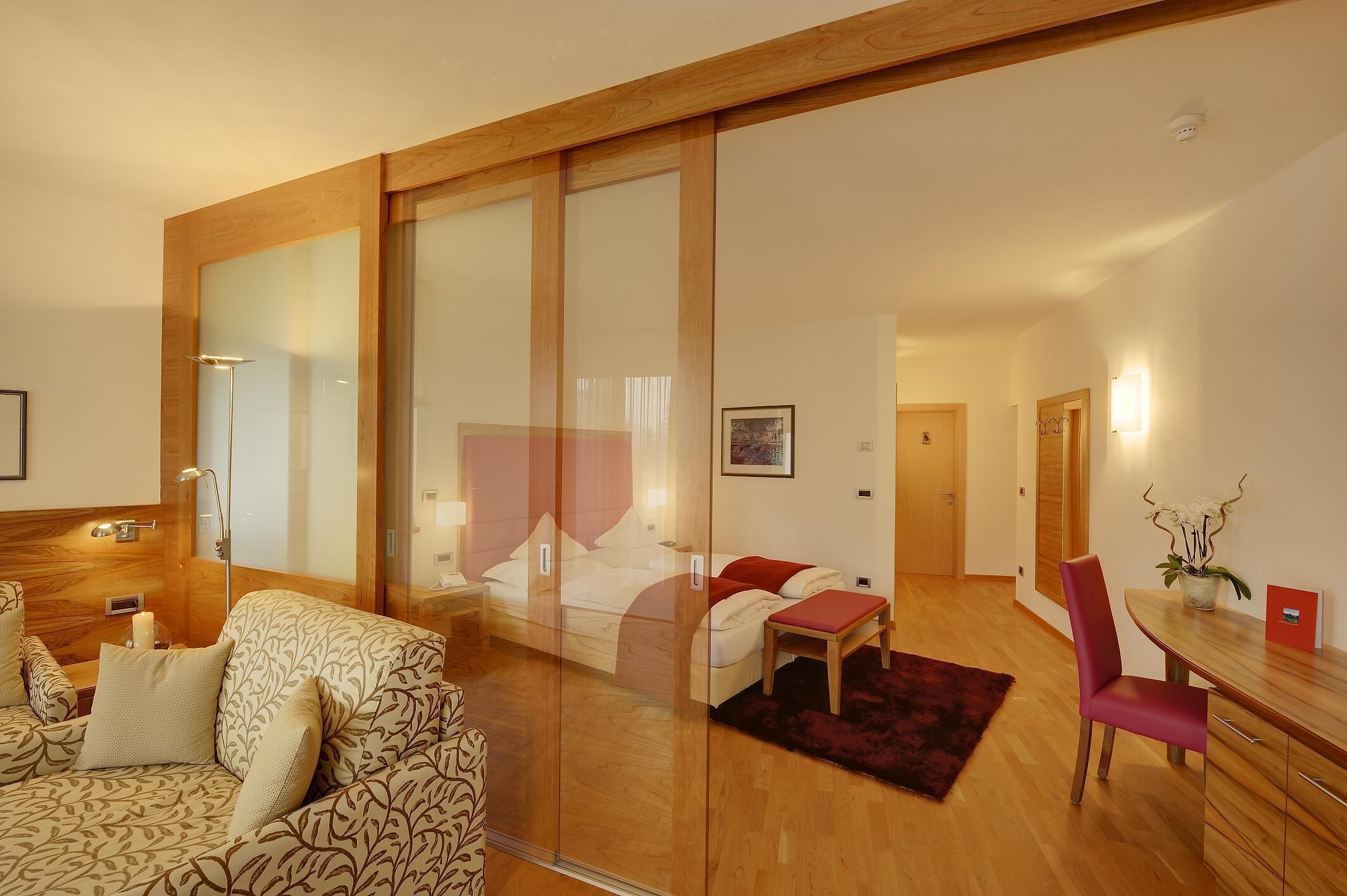 A modern hotel room with a large bed, a sofa, a dining table, and a glass partition.