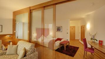 A modern hotel room with a large bed, a sofa, a dining table, and a glass partition.