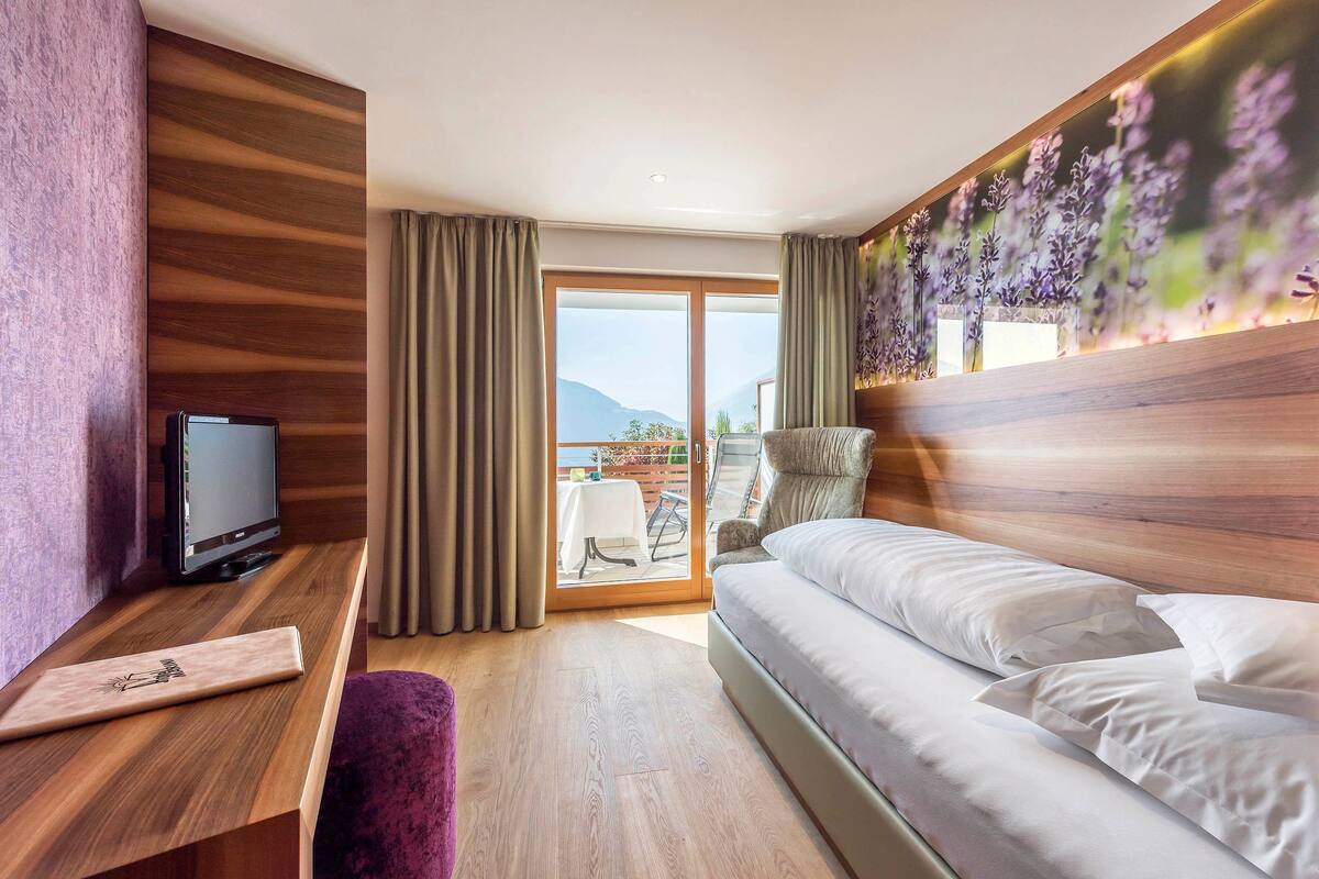 A modern hotel room with a wooden accent wall, a flat-screen TV, a sofa bed, and a balcony with a table and chairs.