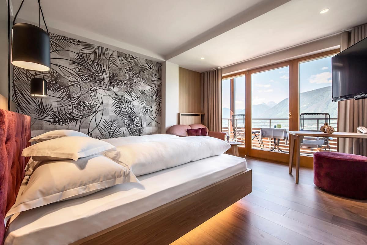 A modern hotel room with a large bed, a balcony with a view of mountains, a TV, and a dining area.