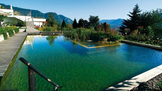 A clear pool with a wooden deck and steps leading into it, surrounded by greenery and mountains in the background. - MARINIs giardino Hotel (Tirolo)