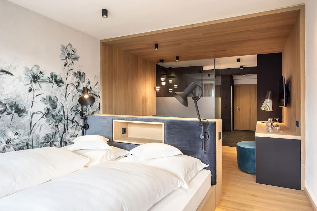 A modern bedroom with a large bed, a bedside table, a desk, and a chair. There is a mirror above the desk and a TV mounted on the wall.