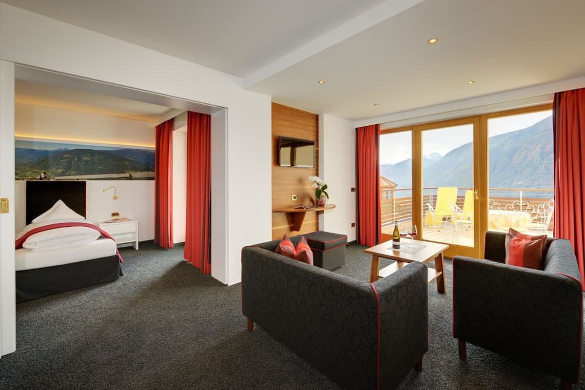 A hotel room with a bed, two sofas, a small table, and a balcony with a mountain view.