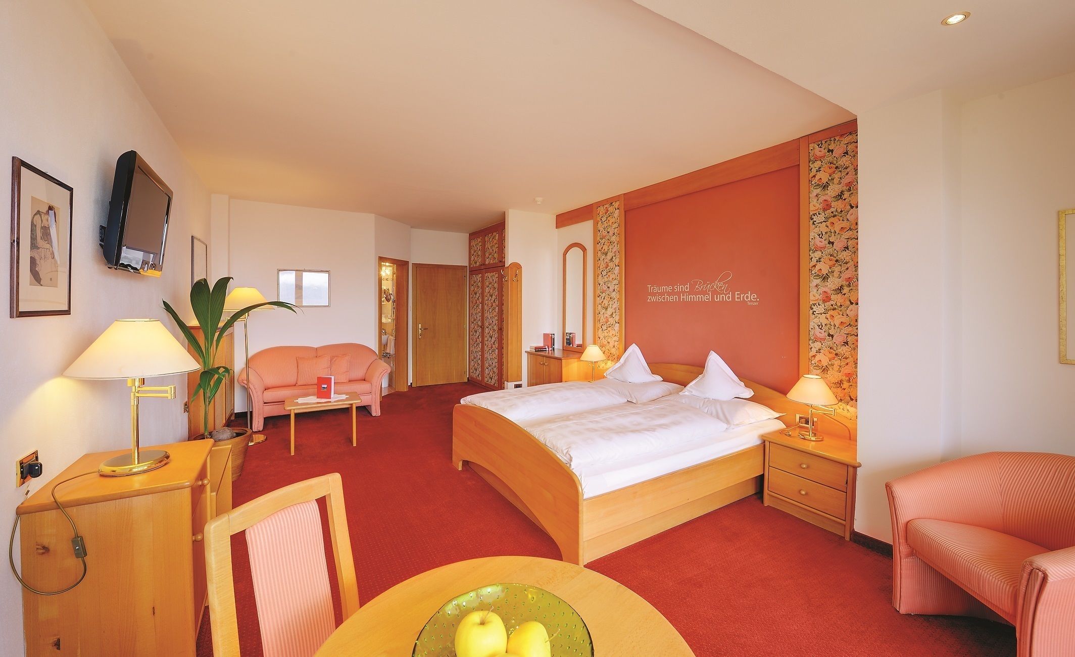 Ett hotellrum med en säng, en soffa, ett skrivbord med en lampa, en stol och ett litet bord med äpplen.
