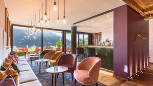A modern lounge area with a sofa, chairs, and tables, a bar counter, and large windows offering a mountain view. - MARINIs giardino Hotel (Tirolo)