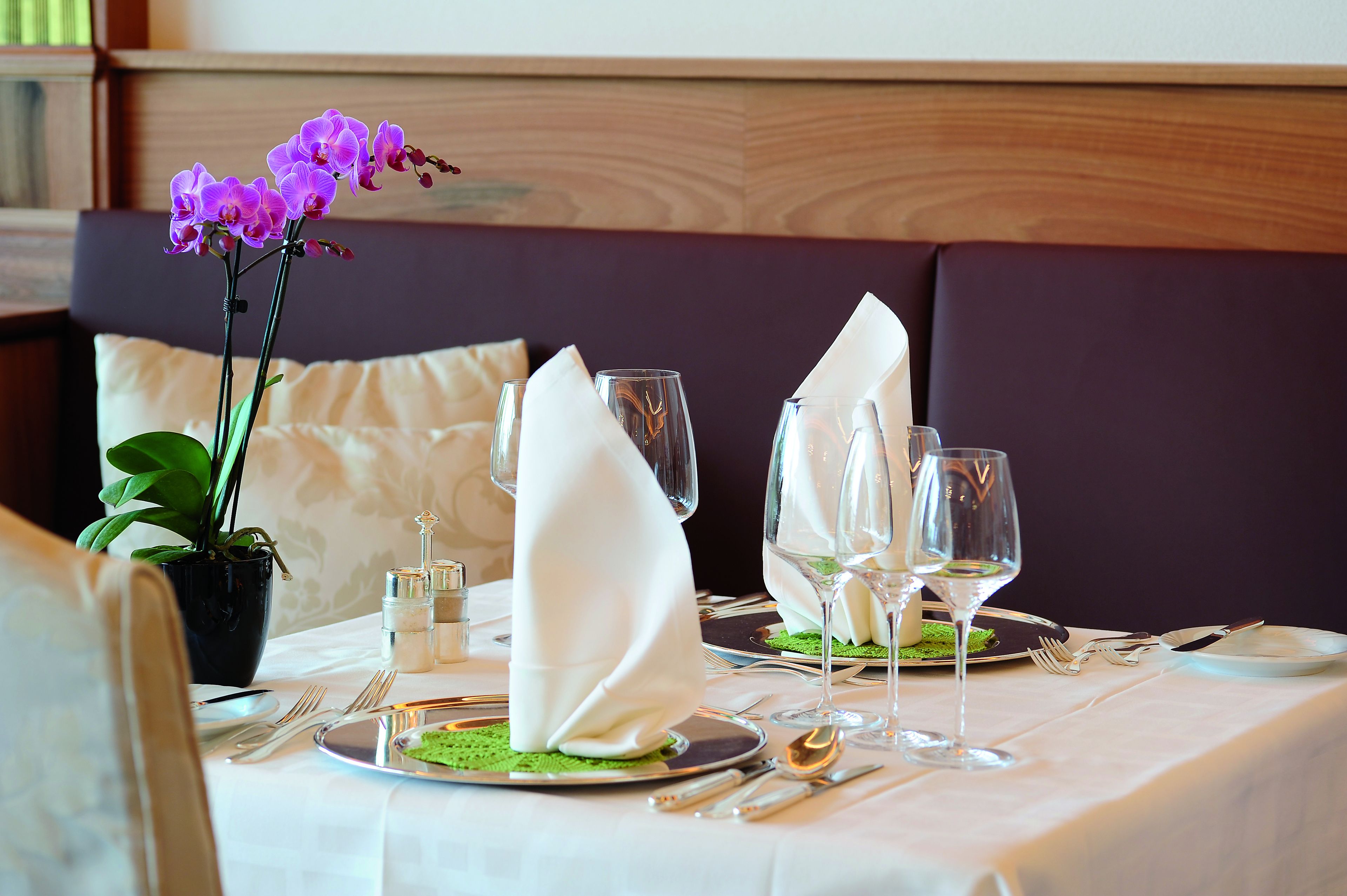 A dining table set with white napkins, wine glasses, and cutlery, with a purple orchid and a potted plant in the background.