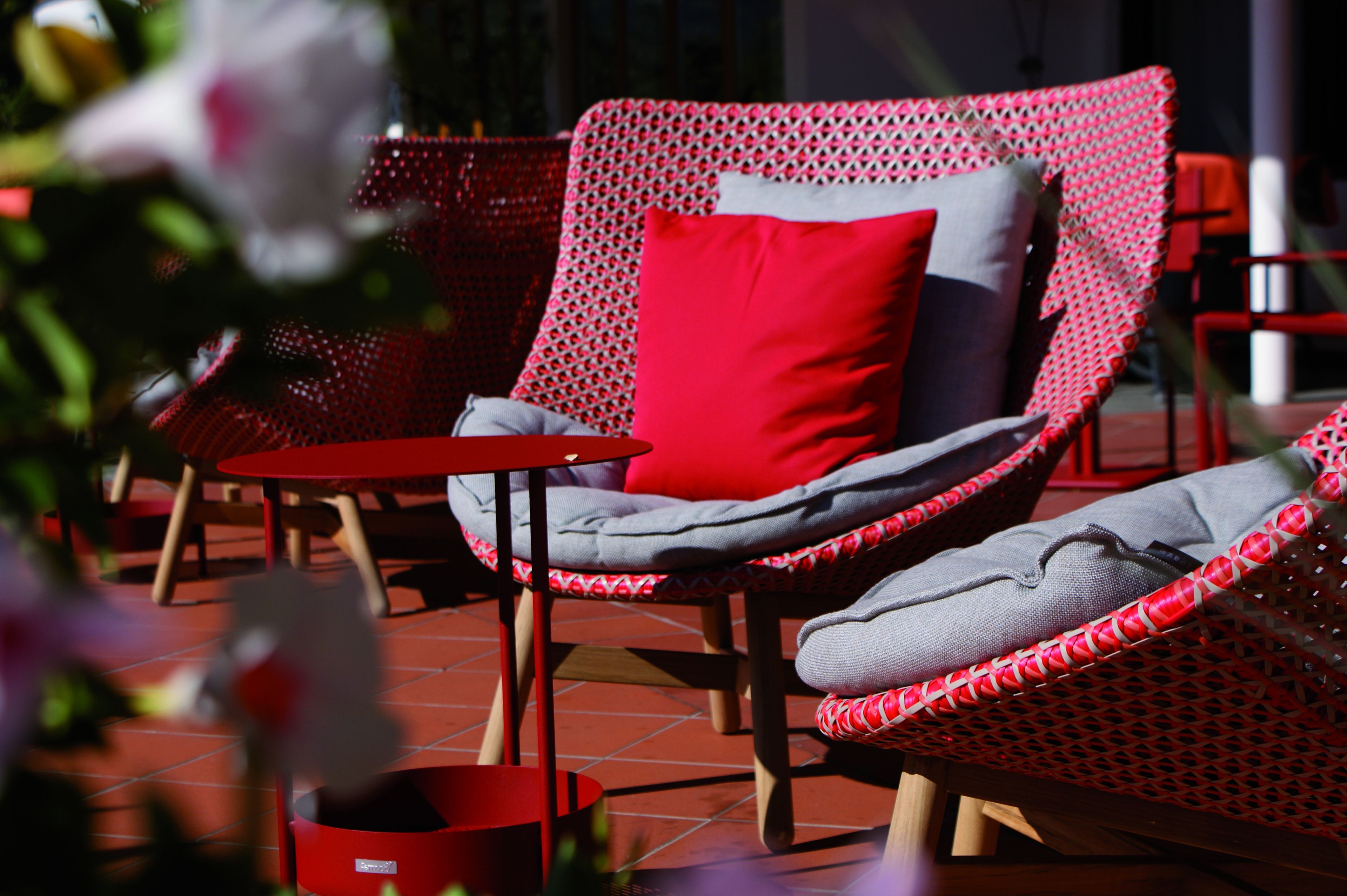 Une chaise en osier rouge, agrémentée d’un coussin gris et d’un oreiller rouge, est placée sur une terrasse carrelée.