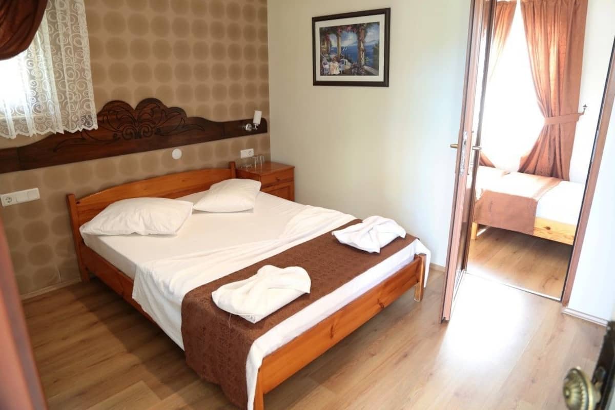 A bedroom with a wooden bed, white linens, a brown throw blanket, a nightstand, a painting on the wall, and a door leading to another room.