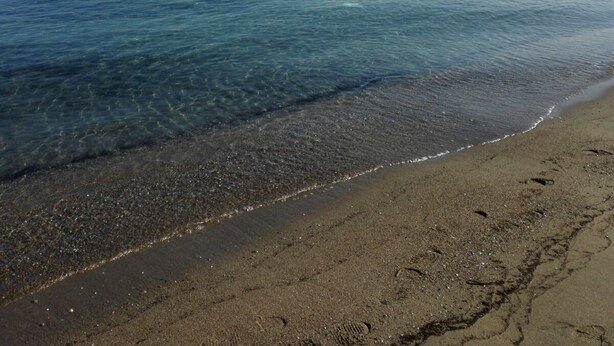 Ein Strand mit klarem Wasser, sandigem Ufer und einer entfernten Küstenlinie mit Gebäuden.