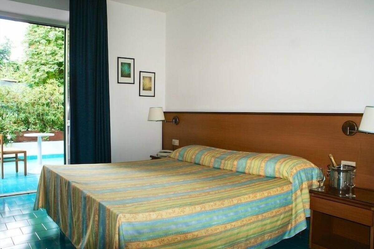 A hotel room with a bed, a desk, a chair, and a window with a view of greenery.