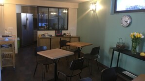 A compact kitchen with a refrigerator, a dining table with chairs, a small wooden table, and a clock on the wall.