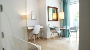 A modern hotel room with a wooden floor, a desk with a chair, a sofa, and a dining area with chairs.