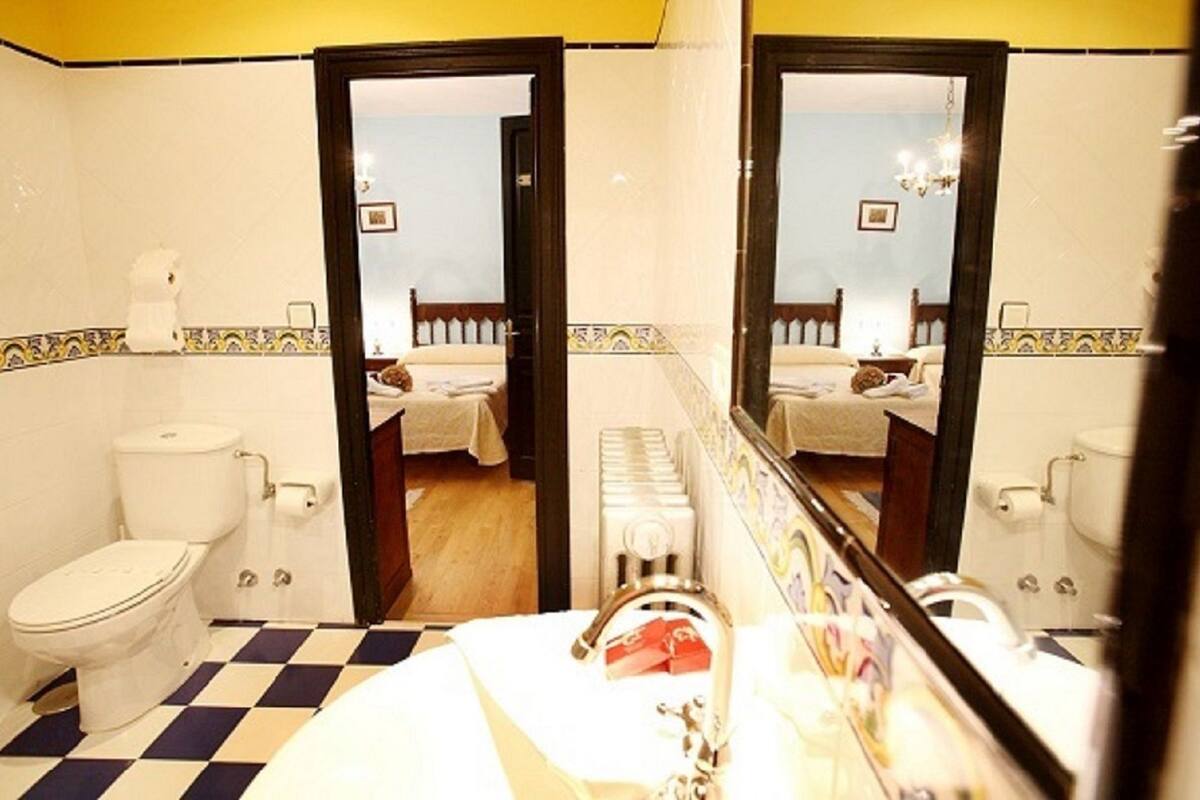 A bathroom with a checkered floor, a toilet, a sink with a mirror, and a bed visible through an open door.
