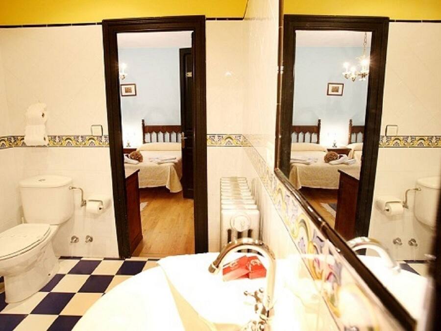A bathroom with a checkered floor, a toilet, a sink with a mirror, and a bed visible through an open door.