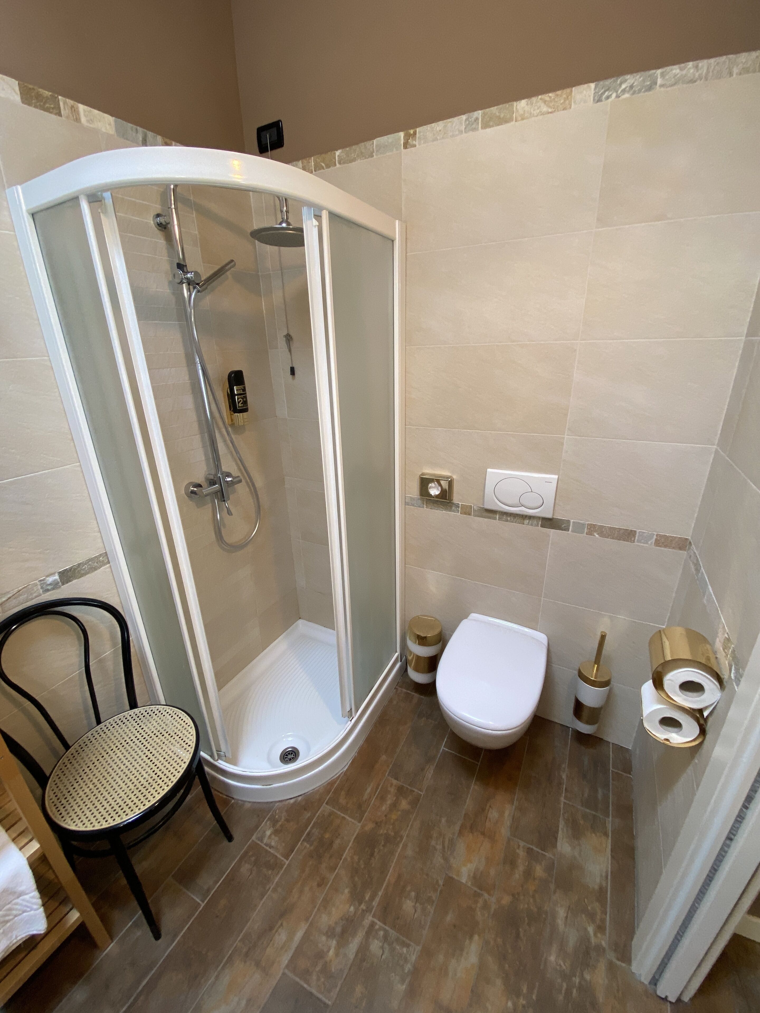 A shower with a glass door, a toilet, and a chair in a bathroom with tiled walls and floors.