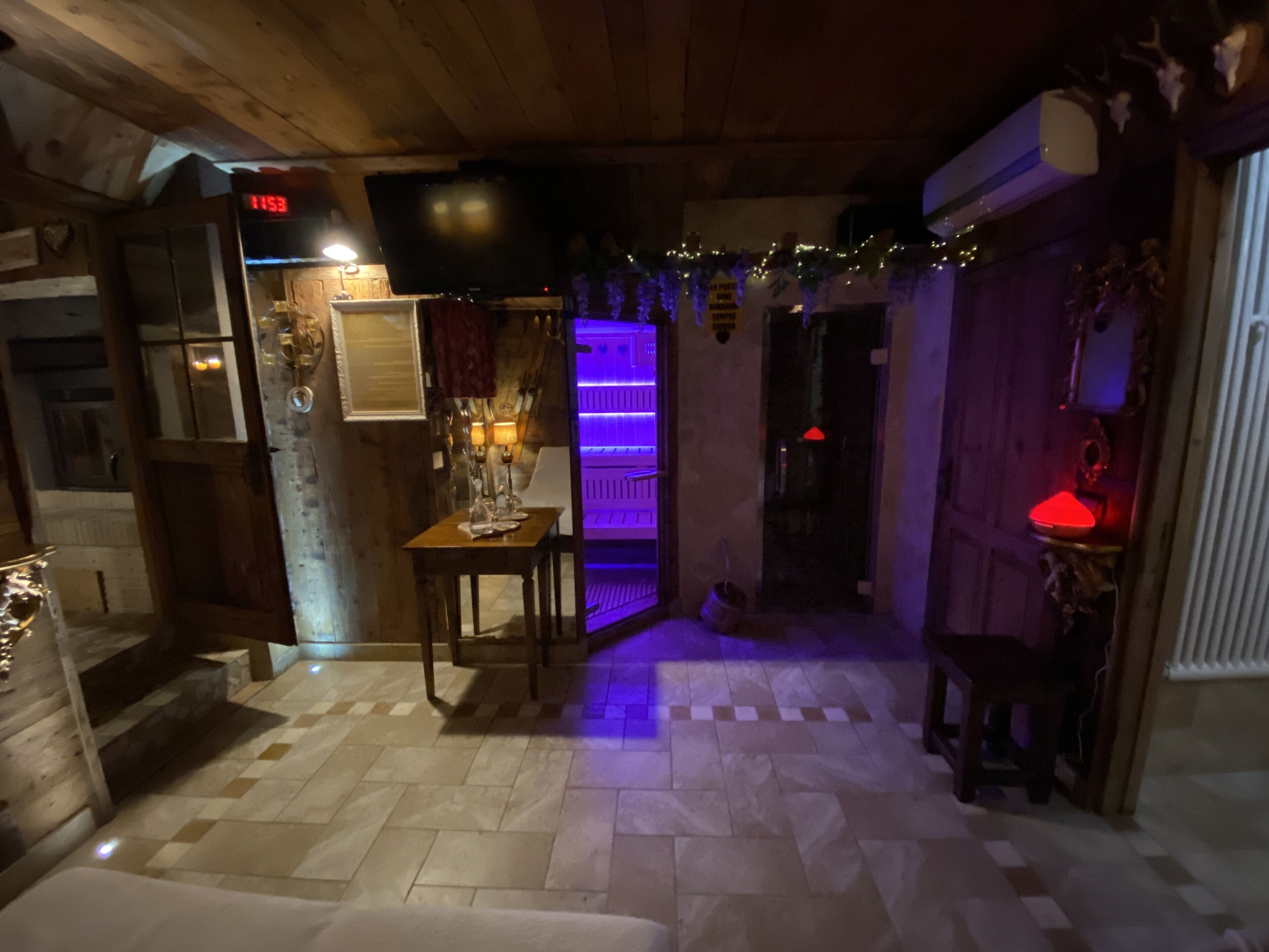Une pièce chaleureuse avec un plafond en bois, une horloge, une table avec des bougies et une étagère illuminée de lumière violette.