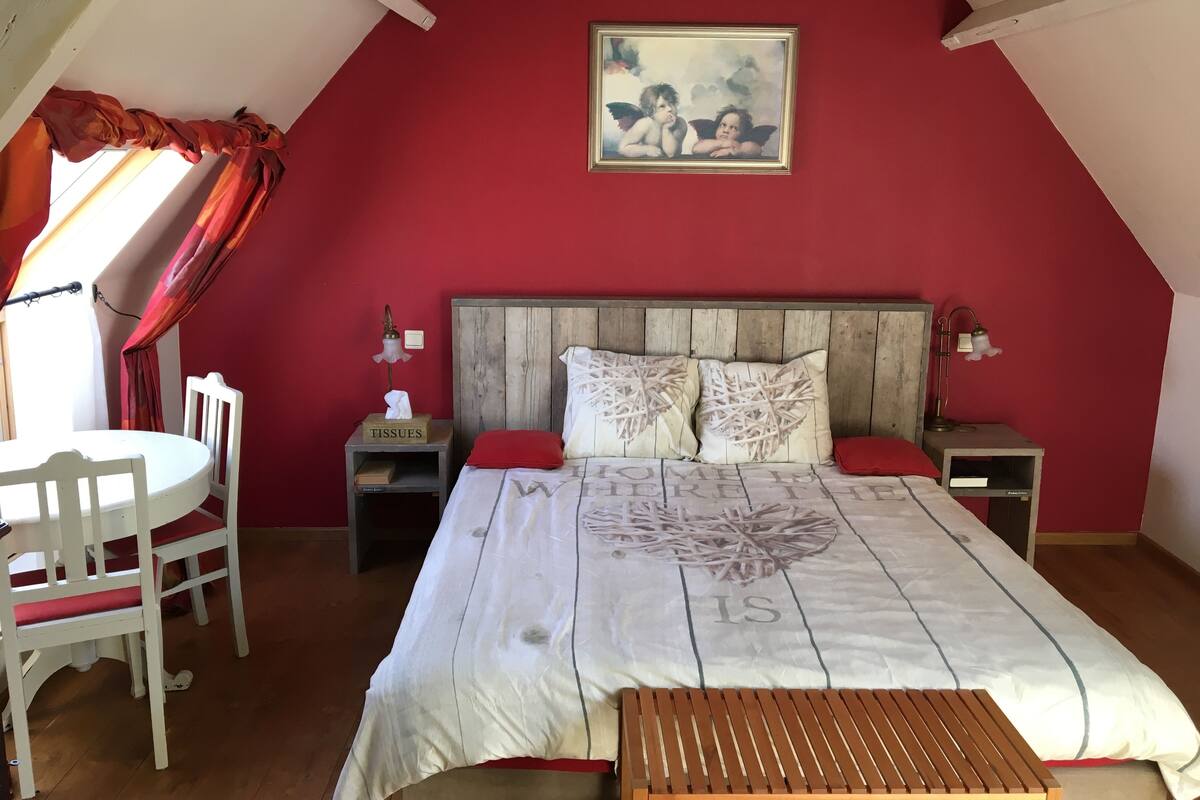 Um quarto com teto inclinado, uma cama com edredom branco, cabeceira de madeira, duas mesas de cabeceira com abajures, uma cadeira vermelha e uma pequena mesa.