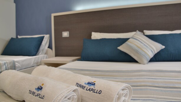 A hotel room with two beds, each with blue and white striped bedding, and a headboard. There are towels with the hotel name on them. - Affittacamere Torre Lapillo (Porto Cesareo)