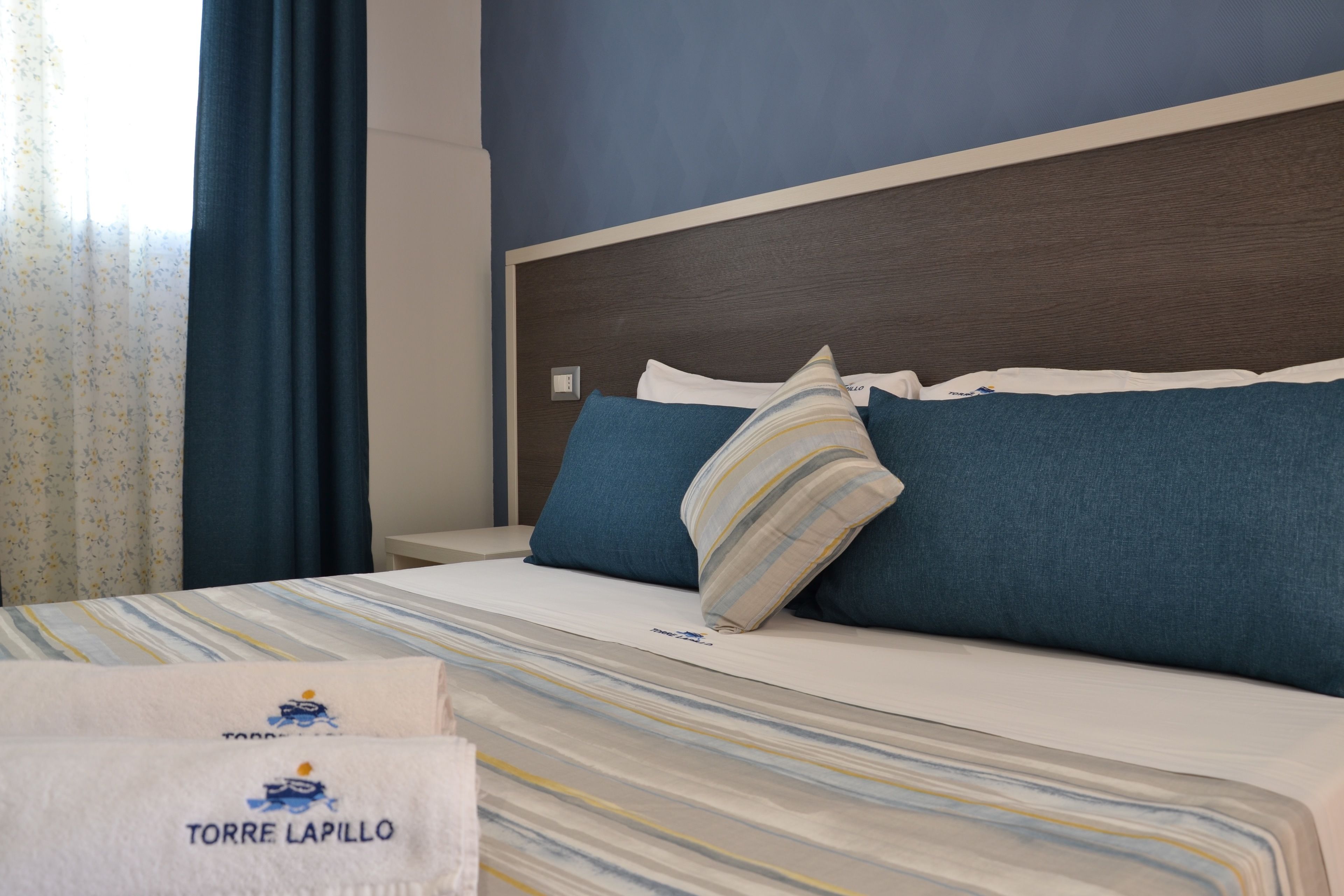 A hotel room with a bed, pillows, a nightstand, and a window with curtains.
