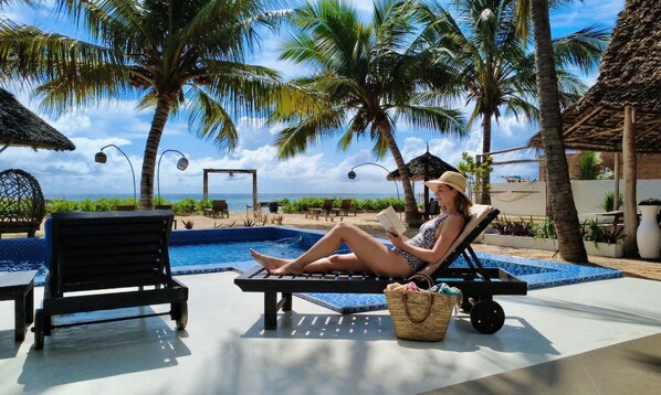 Un espace aménagé au bord de la piscine, avec des chaises longues, une piscine, des palmiers et une vue sur l’océan.