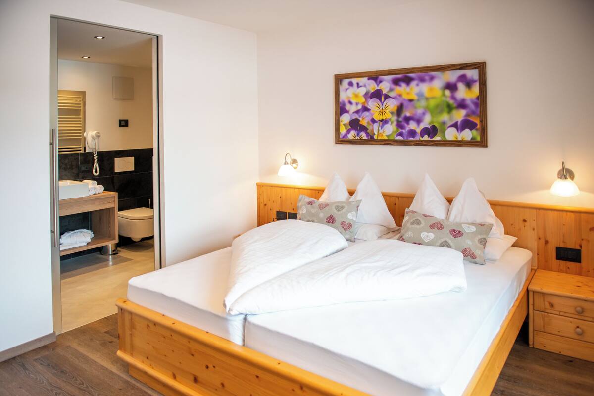 Un dormitorio con cama de madera, ropa de cama blanca, cabecero de madera, lámparas de noche y un cuadro floral en la pared.