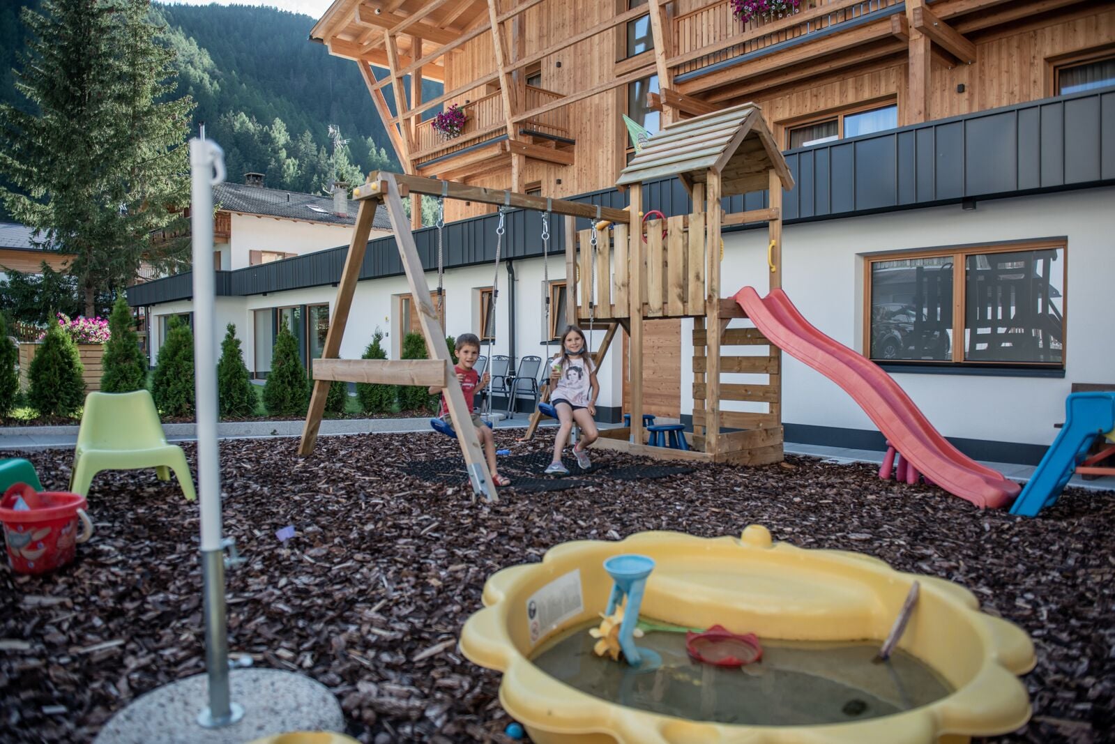 Ein Spielplatz mit Holzspielgeräten, darunter eine Rutsche und ein Kletterbereich, umgeben von Rindenmulch.