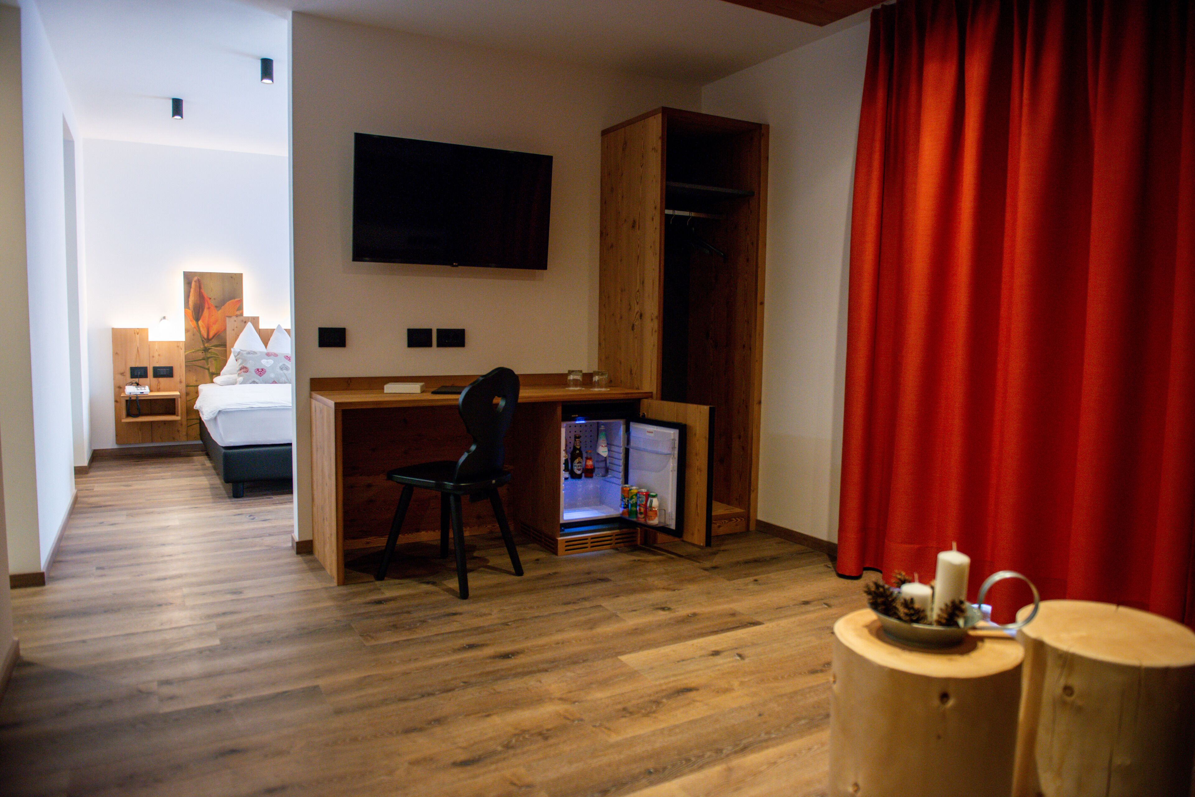 A hotel room with a wooden floor, a bed, a desk with a chair, a television, a wardrobe, and a red curtain.