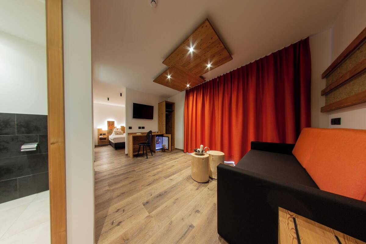 Una sala moderna con suelo de madera, un sofá negro, techo de madera con iluminación empotrada, una cortina roja y un televisor en la pared.