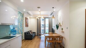 A modern kitchen with white cabinets, a large water dispenser, and a patterned backsplash. There is a dining area with a wooden table and chairs, a gray sofa, and a large window with curtains. - Park Hill No1 Apartment (Hanoi)